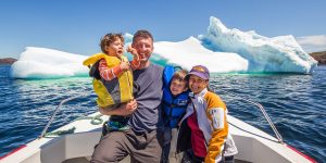 A young family smiles from the bow of a small boat while a large iceberg floats behind them - Icebergs in Twillingate